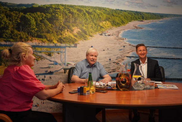 Mężczyzna w szarek koszuli, mężczyzna w ciemnej marynarce i białej koszuli oraz kobieta w fuksjowej bluzce uśmiechnięci siedzą przy okagłym stole. Za nimi, na ścianie - fototapeta przedstawiająca morski brzeg, plażę. Na stole - dokumenty, pieczątki.