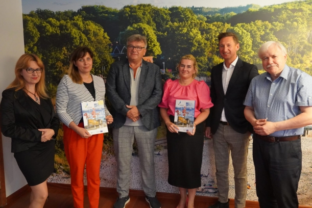 Trzy kobiety i trzech mężczyzn stoją w pomieszczeniu, w którym ścianę zdobi fototapeta przedstawiająca plażę i porośnięty drzewami nadmorski brzeg. Dwie kobiety trzymają teczki z logo Pomorza Zachodniego i zdjęciem kutra rybackiego.