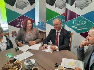 Dwie kobiety i dwóch mężczyżn siedzi przy stole w sali konfernecyjnej; za nimi ustawione są trzy roll-upy z napisem "Darłowska Lokalna Grupa Rybacka"; mężyczna w ciemnym garniturze trzyma długopis, kobieta trzyma pieczątkę nad rozłożonymi dokumentami.