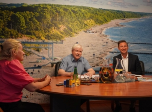 Mężczyzna w szarek koszuli, mężczyzna w ciemnej marynarce i białej koszuli oraz kobieta w fuksjowej bluzce uśmiechnięci siedzą przy okagłym stole. Za nimi, na ścianie - fototapeta przedstawiająca morski brzeg, plażę. Na stole - dokumenty, pieczątki.